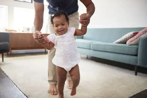 A father helps his baby practice gross motor activities for infants 12-18 months, taking first steps with a smile.
