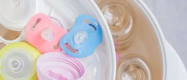 Pink and blue pacifiers sit in a sterilizer with baby bottles. Learn how to sterilize pacifier safely for your baby.