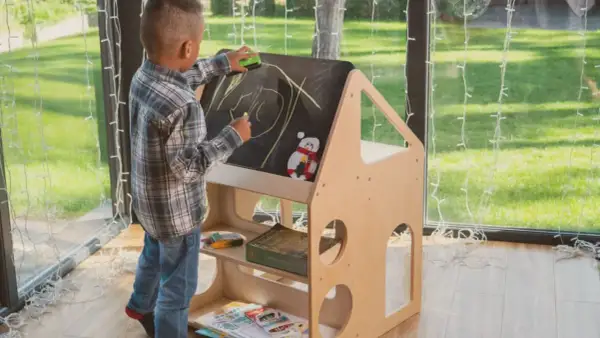 A young boy draws on a chalkboard easel, part of a Montessori backyard playhouse. He's erasing a drawing with a green eraser.