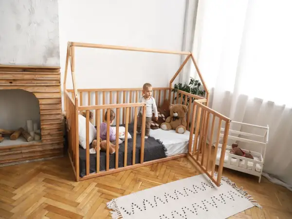 Two children play in a wooden playpen floor bed. One reads a book, the other stands, with a teddy bear nearby.