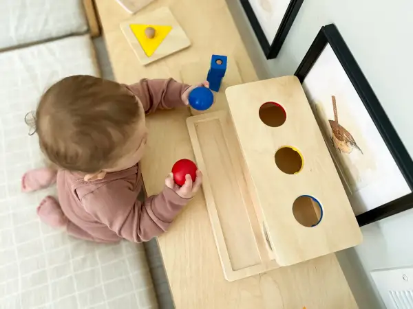 A baby explores Montessori activities for 10 month old baby, holding red and blue balls near a wooden drop box and shape puzzle.