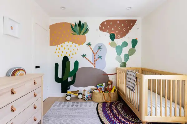 A colorful gender neutral nursery with a desert mural, wooden crib, dresser, and toys.