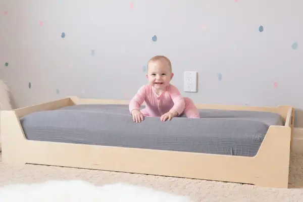 A smiling baby in pink pajamas explores a Montessori bed with a grey mattress. The wooden bed frame is low to the ground, perfect for little adventurers.