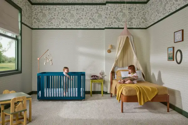 A teal crib offers bold crib color ideas in a nursery with whimsical wallpaper. Two children enjoy the room.