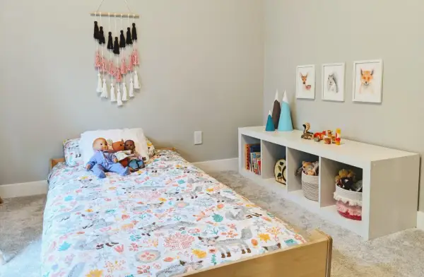 A cozy floor bed setup with a woodland-themed duvet cover, dolls, and a wall hanging. A white cube shelf holds books and toys.