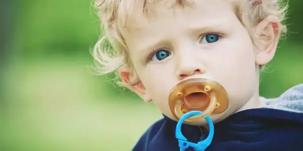 A toddler with blue eyes and blonde hair has a pacifier in their mouth. This image prompts the question: can pacifiers cause cavities?